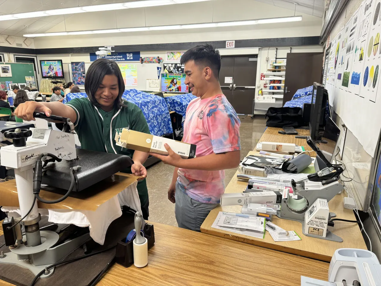 Two Molokaʻi High students working with a heat press and printing equipment in a technology classroom